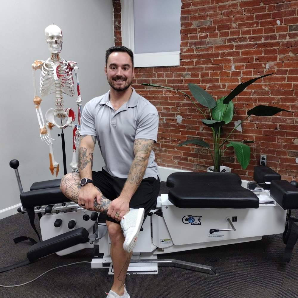 Photo of Dr Kevin Pisle, a Lima, Ohio chiropractor, sitting on his adjustment table next to a skeleton model used for anatomical demonstration.